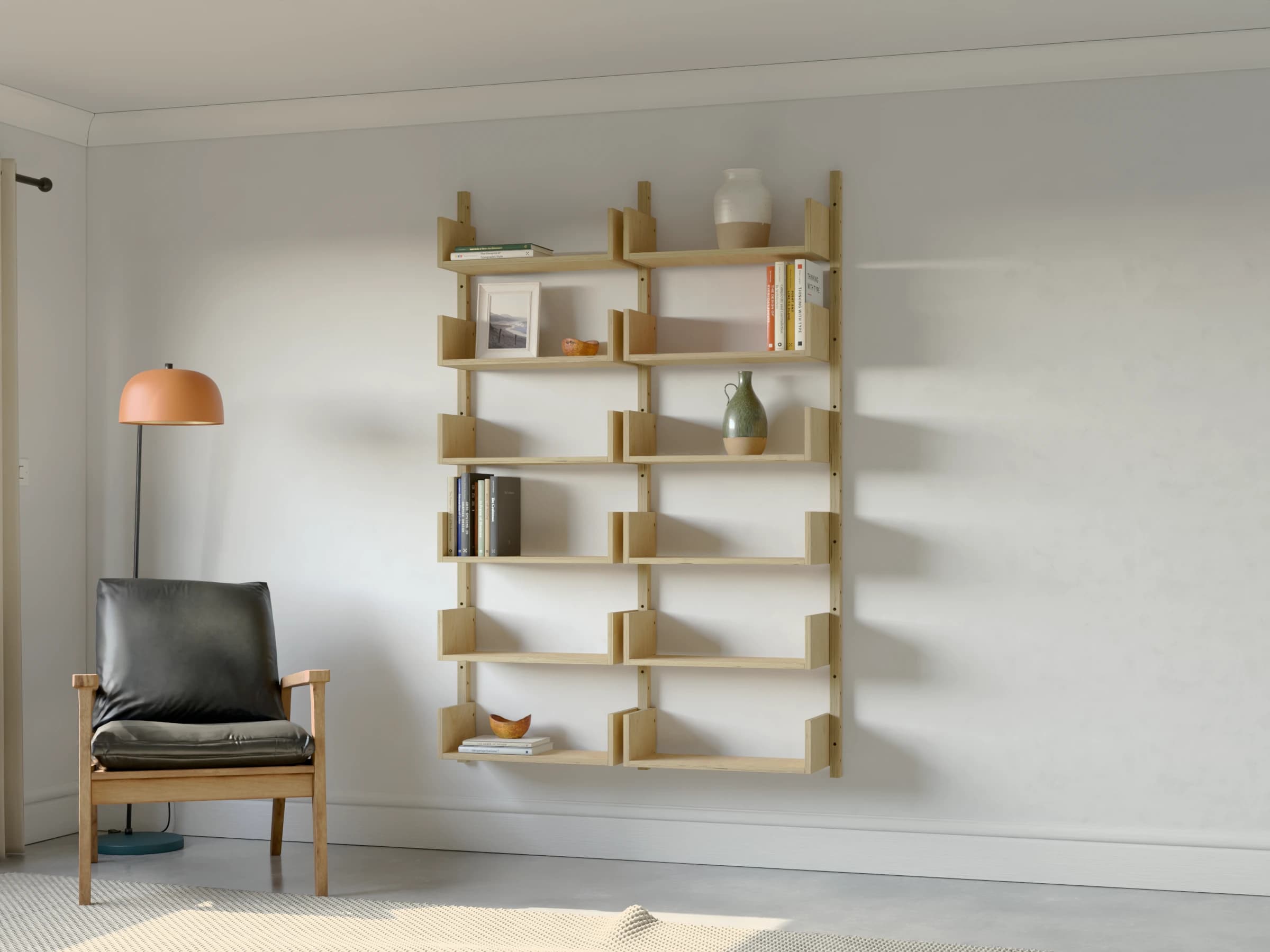 Two bays of birch plywood shelving with six bookended shelves in each bay, styled with books and ceramics beside an armchair and a floor lamp