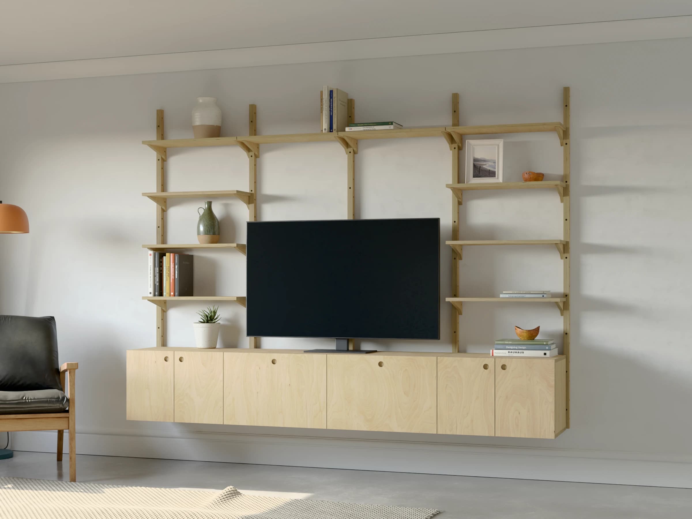 A four-bay birch plywood media wall with a television standing on the cabinets: cupboards in the outer bays, deep drawers in the middle and shelves above, beside an armchair and a floor lamp