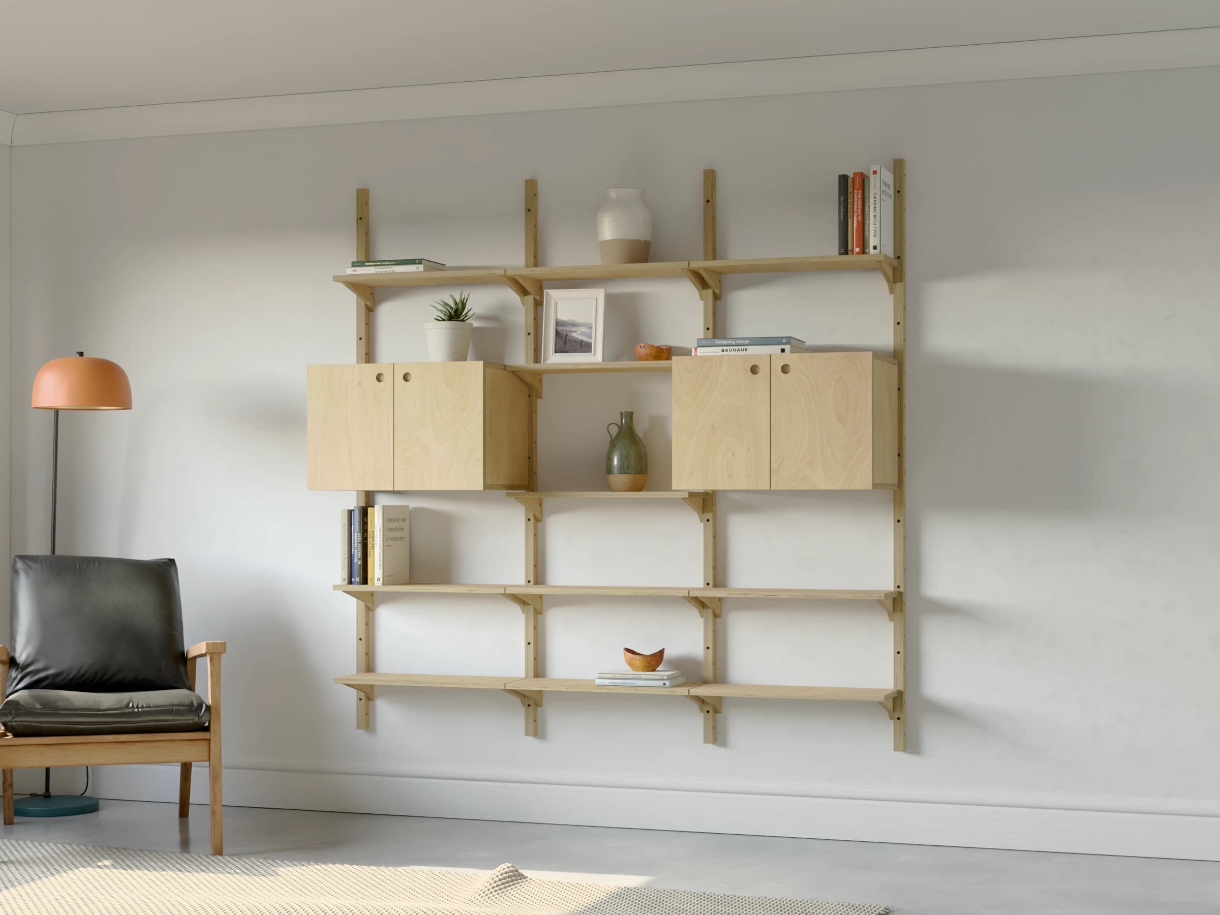 Three bays of birch plywood shelving with two-door cupboards at eye level in the outer bays and open shelves in the middle, styled with books and ceramics beside an armchair and a floor lamp